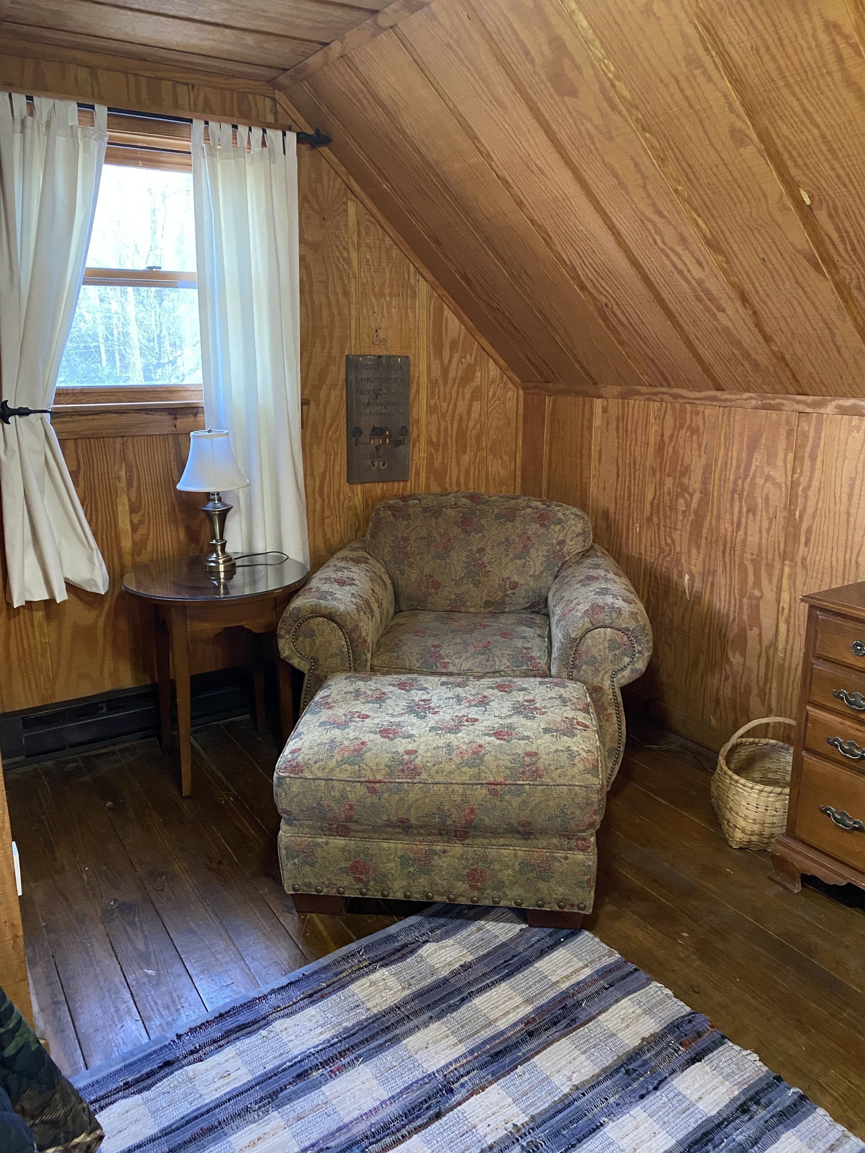 A cozy attic room with wooden walls, a floral-patterned armchair, a small wooden table with a lamp, and a wooden dresser.