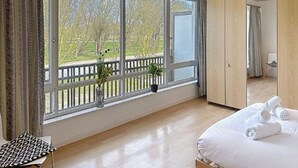 Een slaapkamer met een groot raam, houten vloeren, een bed met witte lakens en uitzicht op bomen en een hek.