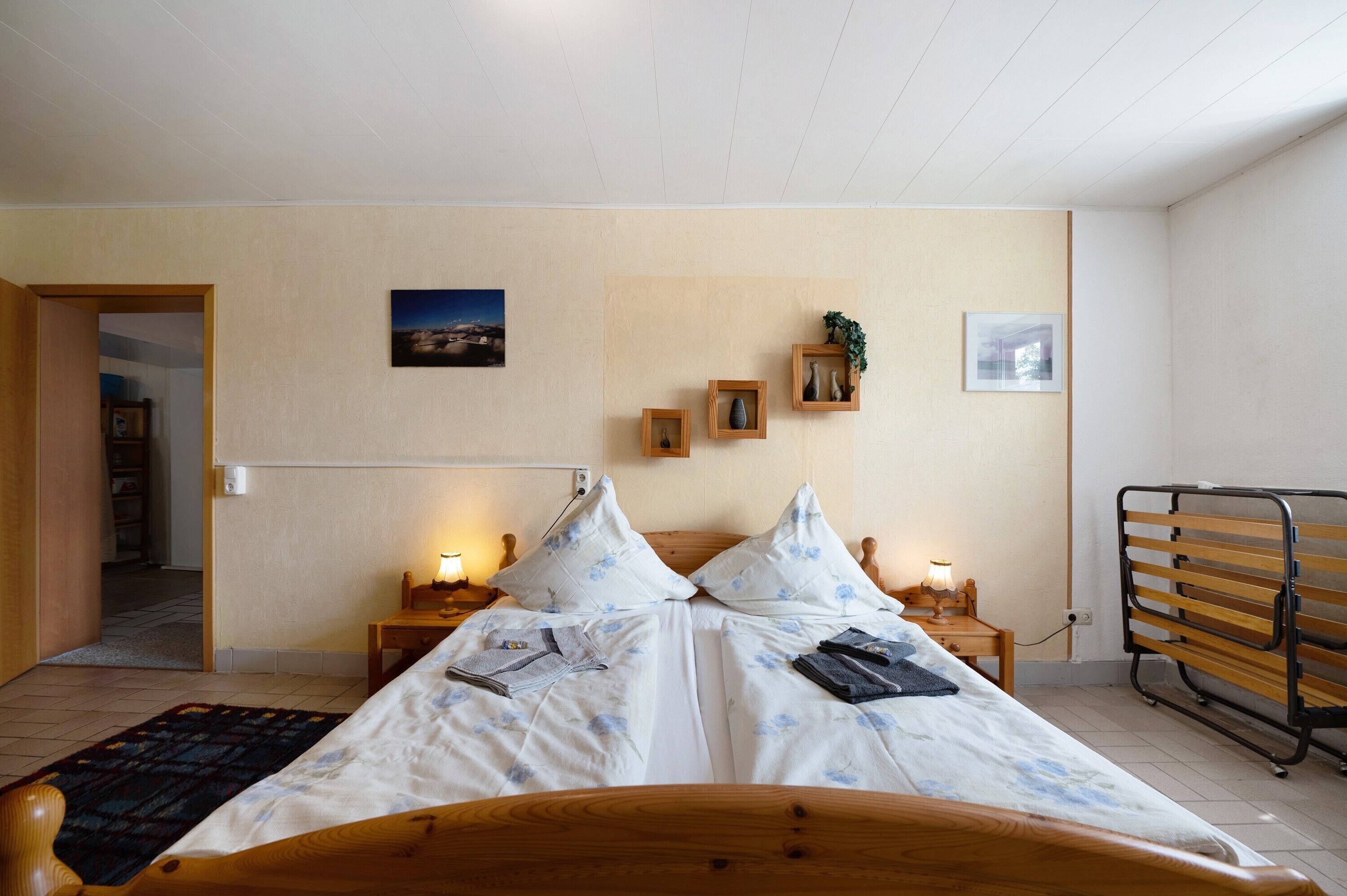 A double bed with a wooden headboard, two bedside tables with lamps, a wooden bench, and a view of a room with a door and a wall-mounted picture.
