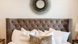 Ein Bett mit einer gepolsterten Kopfstütze, weißen und gemusterten Kissen sowie einem runden Spiegel an der Wand.
