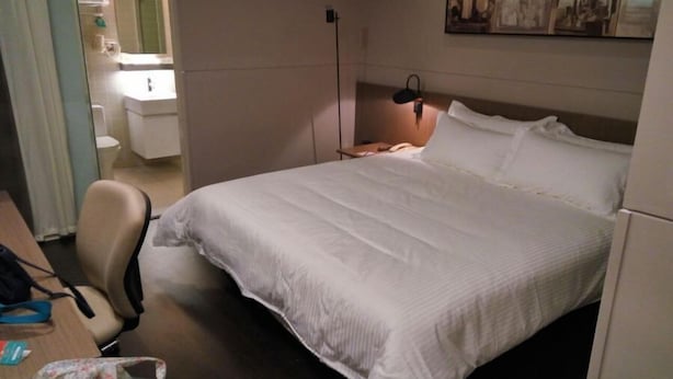 A hotel room with a large bed, a desk with a chair, a bedside table with a lamp, and a bathroom visible through an open door. - Jinjiang Inn Select (Shanghai)
