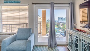 A room with a sliding glass door leading to a balcony with a pool view, a blue armchair, and a cabinet with decorative items. - Coastal Comfort at Beach Barn - Pet Friendly! (Galveston)