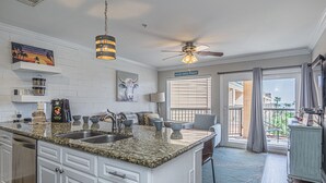 A spacious kitchen with granite countertops, white cabinetry, and a large island. There is a ceiling fan, a window with blinds, and a view of the outdoors. - Coastal Comfort at Beach Barn - Pet Friendly! (Galveston)