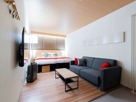 Una habitación de hotel moderna con cama, sofá gris, suelo de madera y televisor de pantalla plana.