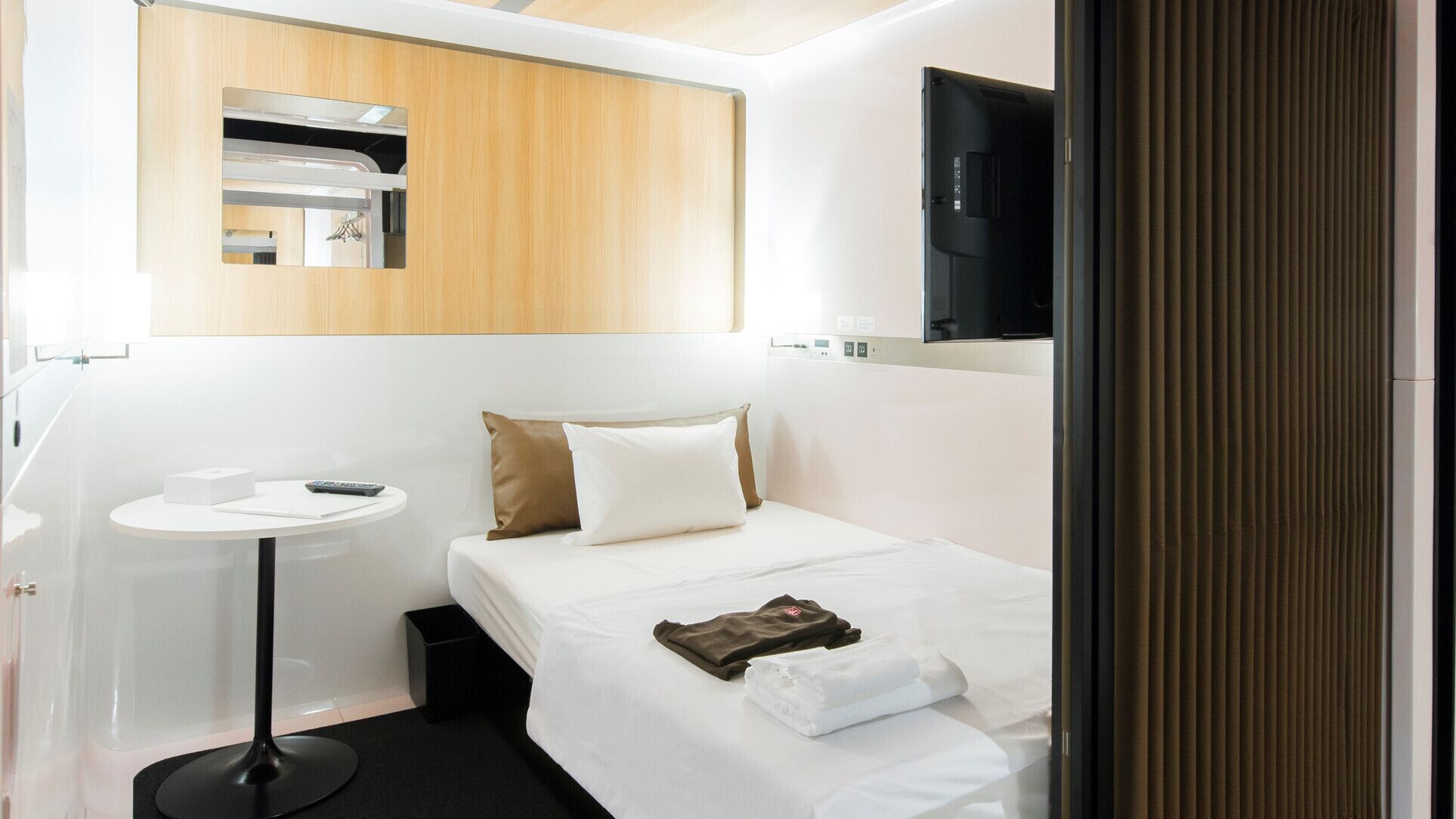 Una habitación de hotel con cama, mesita de noche, una pequeña mesa redonda, espejo y televisión montada en la pared.