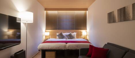 A modern hotel room with a large bed, a flat-screen TV, a sofa, and a window with blinds.