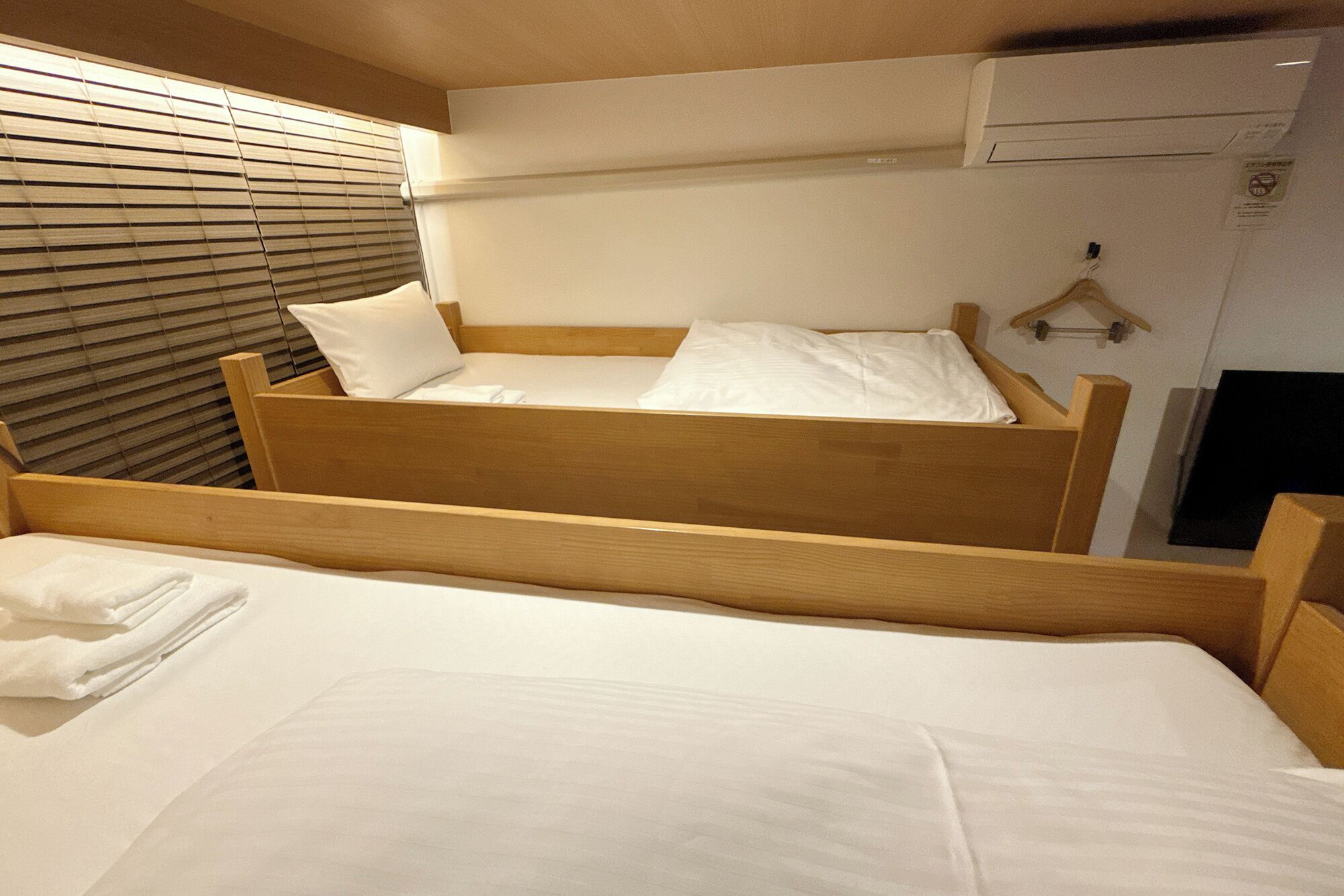 A single bed with a wooden frame and white bedding, a window with blinds, and a wall-mounted air conditioning unit.