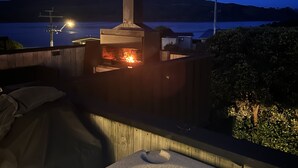 A hot tub with a stone edge and a fire feature, overlooking a body of water with hills in the background. - Raglan Beach House , Stunning Ocean and Harbour views out at  Ocean Beach (Raglan)