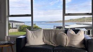 A black leather sofa with white cushions, a round wooden side table, and a beige patterned rug. - Raglan Beach House , Stunning Ocean and Harbour views out at  Ocean Beach (Raglan)