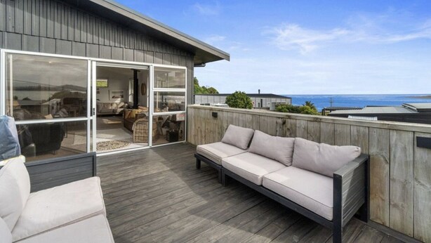 A modern deck with a wooden floor, a beige sofa, and a glass door leading to an interior space. - Raglan Beach House , Stunning Ocean and Harbour views out at  Ocean Beach (Raglan)