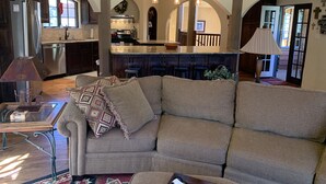 A cozy living room with a sofa, a coffee table, and a lamp. There is a kitchen area with a refrigerator, a sink, and a stove. The room has arched ceilings and pendant lights.