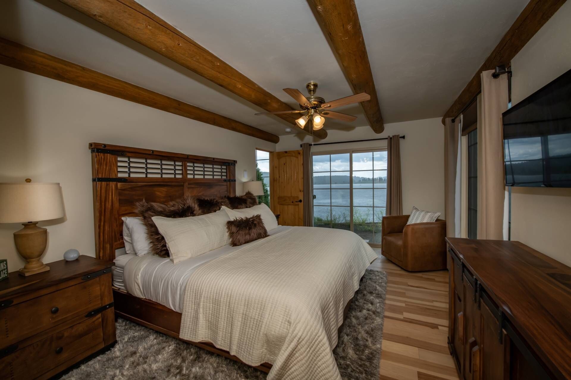 Une chambre à coucher avec une tête de lit en bois, un lit recouvert de linge de lit blanc, une table de chevet en bois avec une lampe, une commode et une vue sur un lac, visible à travers de grandes fenêtres.
