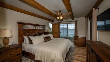 Une chambre à coucher avec une tête de lit en bois, un lit recouvert de linge de lit blanc, une table de chevet en bois avec une lampe, une commode et une vue sur un lac, visible à travers de grandes fenêtres.