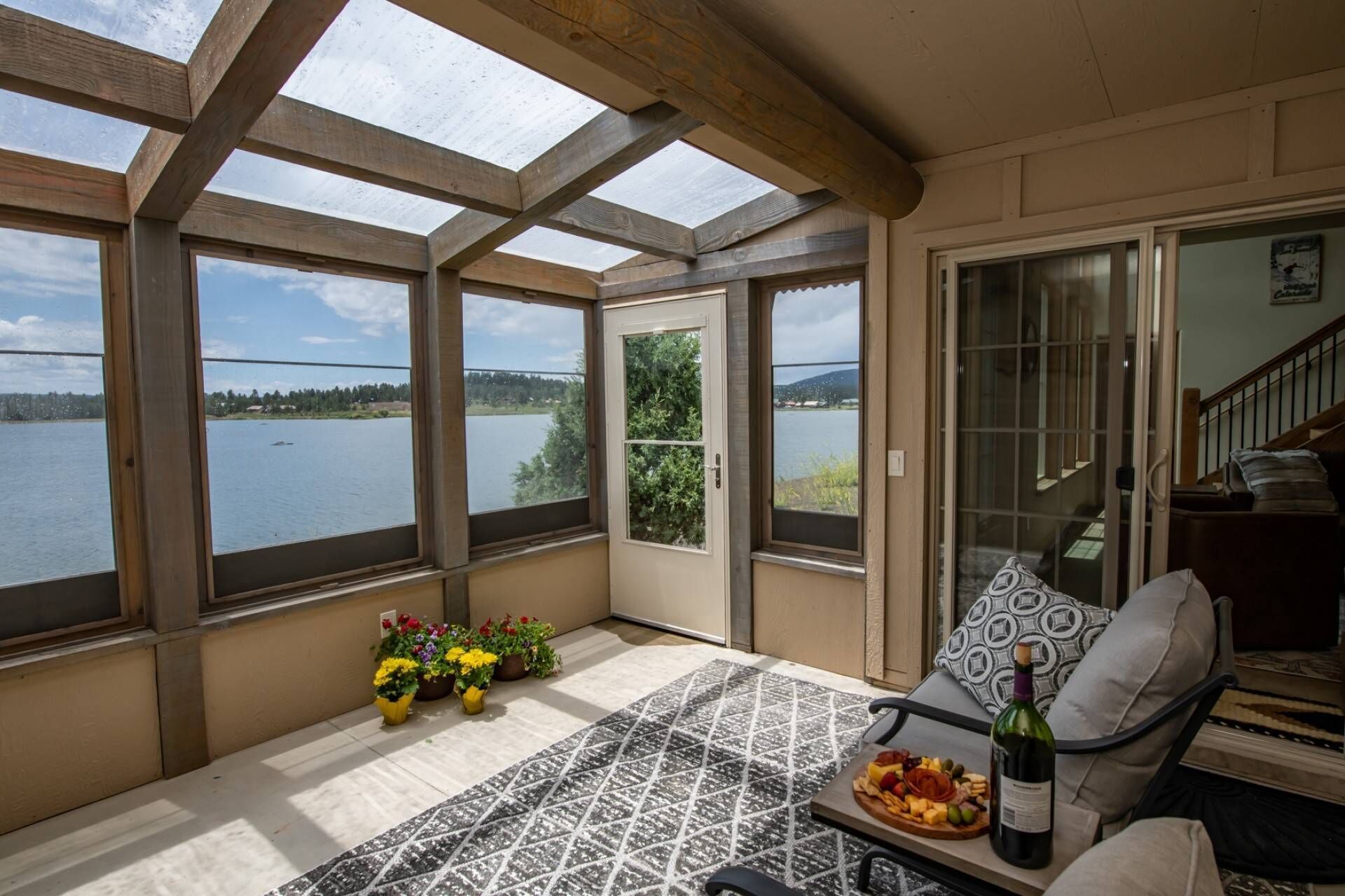 Une pièce baignée de lumière avec vue sur un lac, une table à manger avec une bouteille de vin, et un canapé agrémenté d’un coussin à motifs.