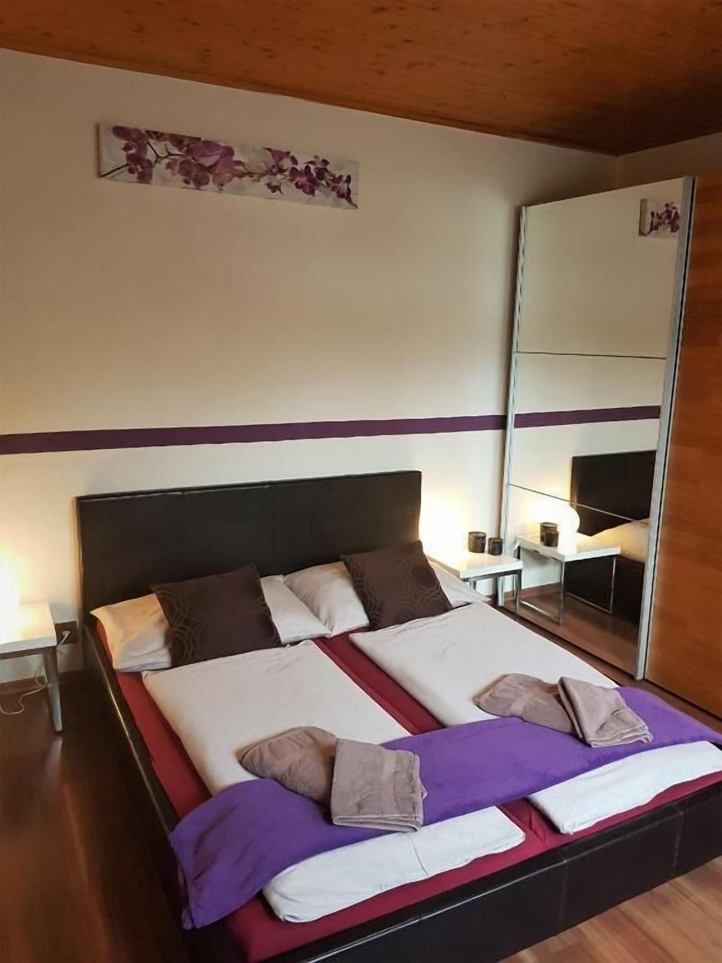 Ein ordentlich bezogenes Bett mit Kopfteil, ein Nachttisch mit Lampe und ein großer Spiegel an der Wand.