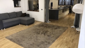 A modern living room with a grey sectional sofa, a large grey rug, wooden flooring, a kitchen area with a grey countertop, and a dining table with chairs. - Pension Günther (Graz)