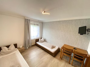 Une pièce de taille modeste comprenant un lit, un petit canapé, une table à manger avec des chaises, un téléviseur fixé au mur et une fenêtre avec des rideaux.