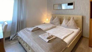 A neatly made bed with a herringbone-patterned throw, a bedside table with a lamp, and a view of a window with curtains. - Pension Günther (Graz)