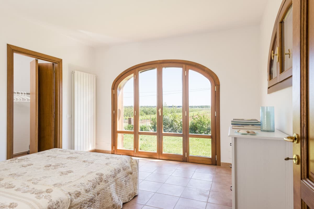Une chambre dotée d’une grande fenêtre en arc de cercle, d’un lit recouvert d’une couvre-lit à motifs floraux, d’une armoire blanche et offrant une vue sur la verdure environnante.