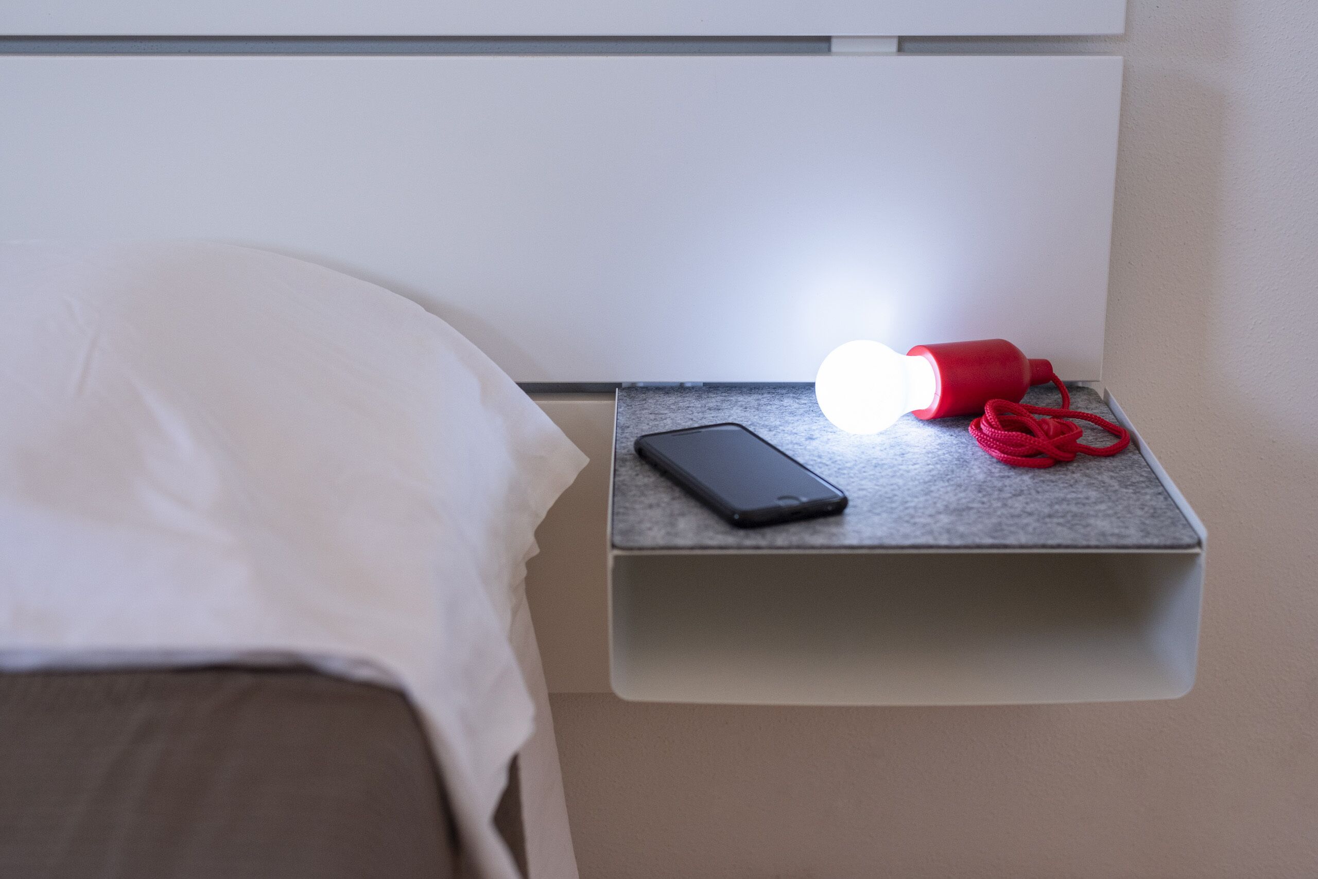 Uma cama com len çol branco, uma mesa de cabeceira com um smartphone e um abajur vermelho, e uma parede com interruptor de luz.