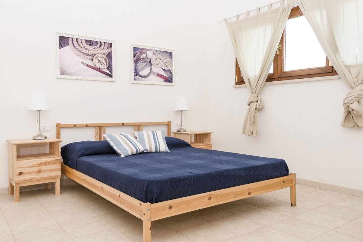 Un lit simple avec un cadre en bois, deux tables de chevet, deux lampes fixées au mur et deux tableaux encadrés.
