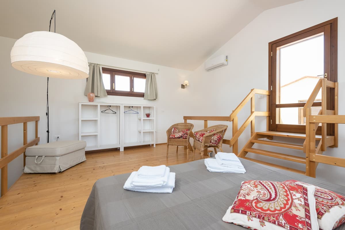 Une chambre à coucher avec un lit, un luminaire suspendu, une fenêtre avec des rideaux, une chaise en bois, une armoire blanche et un escalier en bois.