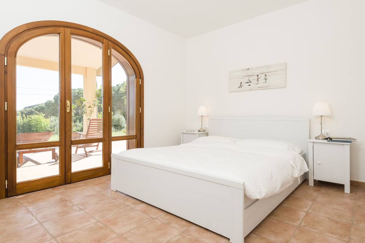 Une chambre avec un grand lit, deux tables de chevet avec des lampes, une porte en bois donnant sur un balcon et un tableau accroché au mur.