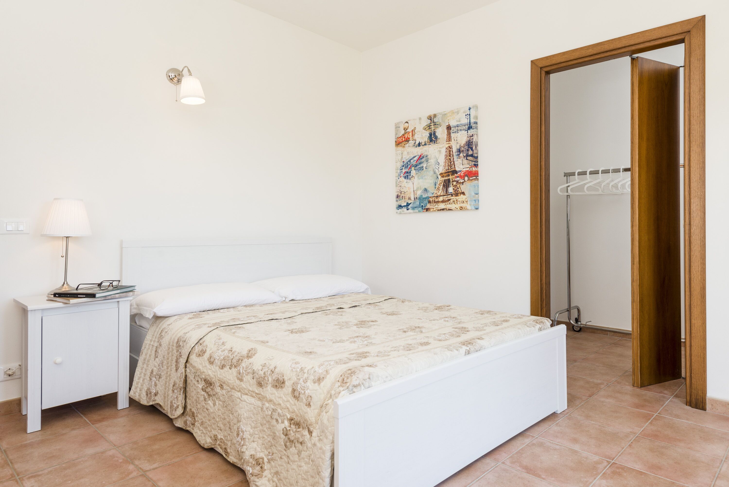 A bedroom with a bed, nightstand, lamp, and a painting of Paris.