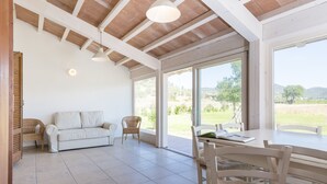 A spacious room with a high ceiling, a white sofa, wicker chairs, a dining table with chairs, and large windows offering a view of the outdoors. - Agriturismo Terre di Castiglione (Castiglione della Pescaia)