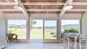 A spacious room with a large sliding glass door leading to a view of a grassy landscape and hills. There is a white dining table with chairs and a wicker chair. - Agriturismo Terre di Castiglione (Castiglione della Pescaia)