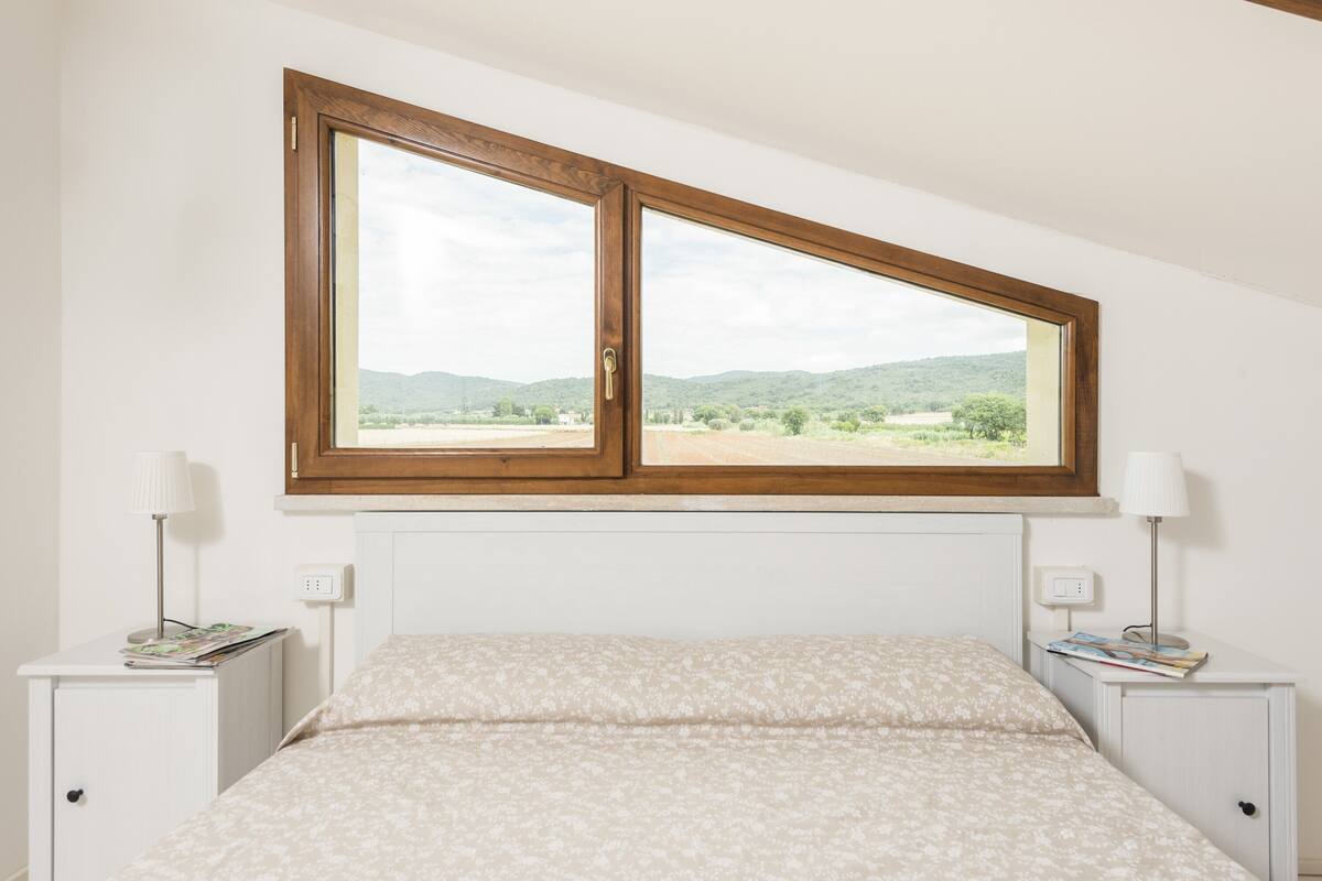 Une chambre à coucher avec un lit, une fenêtre donnant sur des collines, deux tables de chevet avec des lampes et un mur blanc.