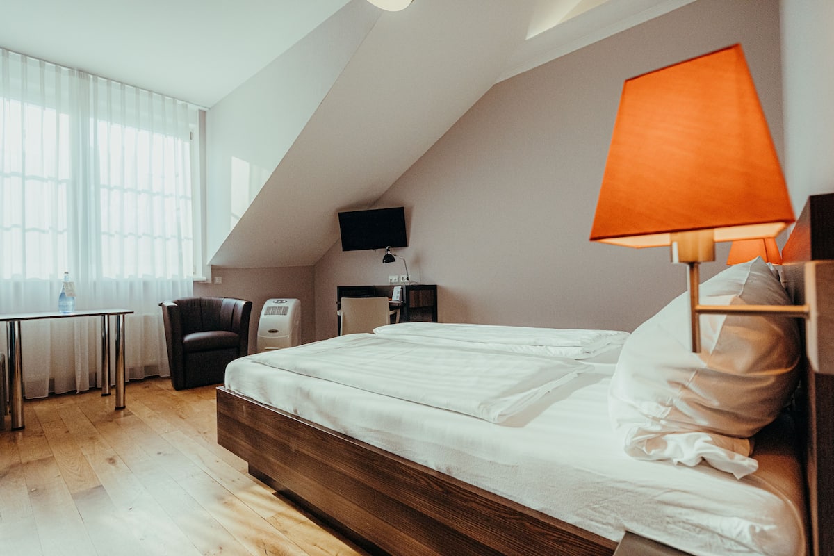 Moderni hotellihuone, jossa on puinen sänky, työpöytä tuolilla, taulu-tv ja suuri ikkuna, jossa on läpikuultavat verhot.