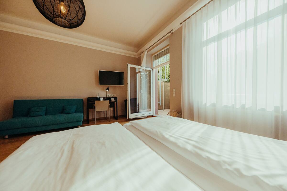 Hotellihuone, jossa on sänky, vihreä sohva, työpöytä tuolilla, televisio ja suuri ikkuna, jossa on läpikuultavat verhot.