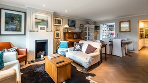 A spacious living room with a fireplace, a dining area, and a kitchen. There are multiple framed artworks on the walls and a bookshelf filled with books. - A Hidden Mews (London)
