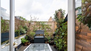 A small, enclosed outdoor space with a glass table, a black chair, and various potted plants. - A Hidden Mews (London)