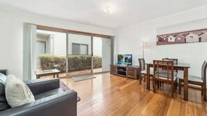 A spacious living room with a wooden floor, a black sofa, a dining table with chairs, and a television. - Secret Dream Location with WIFI and Wine (Rye)