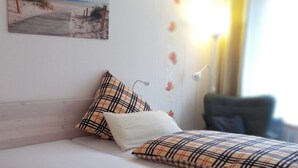 A bedroom with a bed, pillows, a wall-mounted lamp, a painting of a beach, and a window with curtains. - Falkenhof (Westensee)