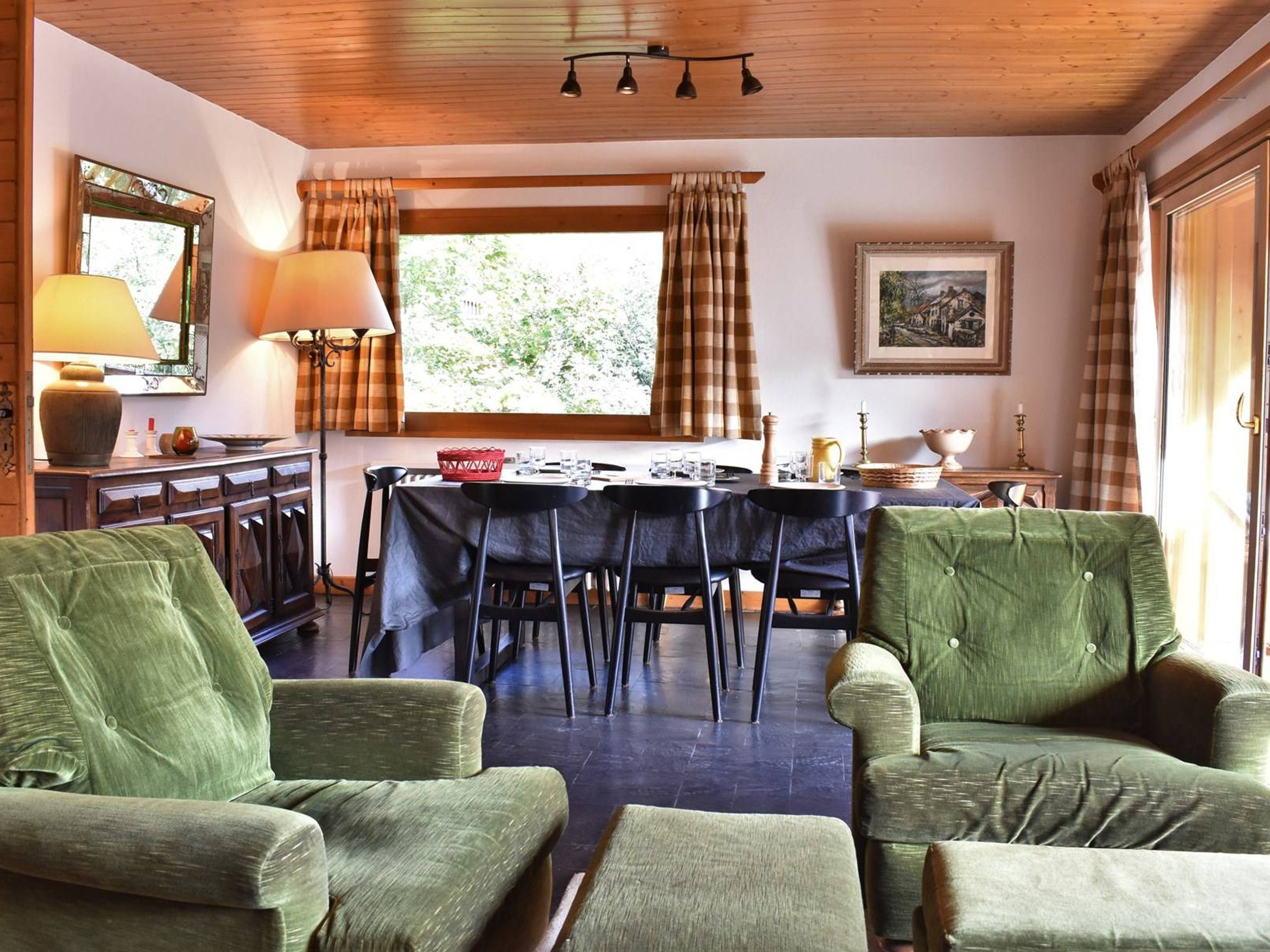 A dining area with a table set for four, surrounded by green upholstered chairs, a wooden ceiling, and a window with curtains.