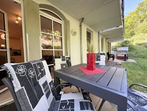 Une terrasse couverte avec une table et des chaises, un vase rouge orné de plantes et une vue sur le jardin.