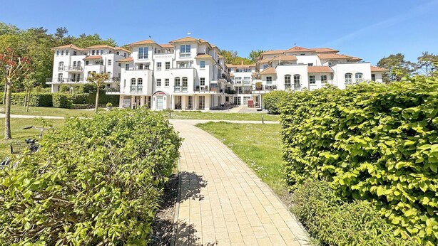 Un edifici gran de diversos pisos amb una entrada central, envoltat per jardins ben cuidats i un camí pavimentat.