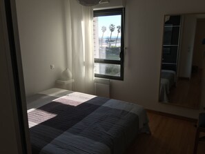 A bedroom with a large window, a bed with a striped bedspread, a bedside table with a lamp, and a mirror on the wall.