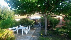 Een terras met een tafel en stoelen, omgeven door groen en een huis op de achtergrond.