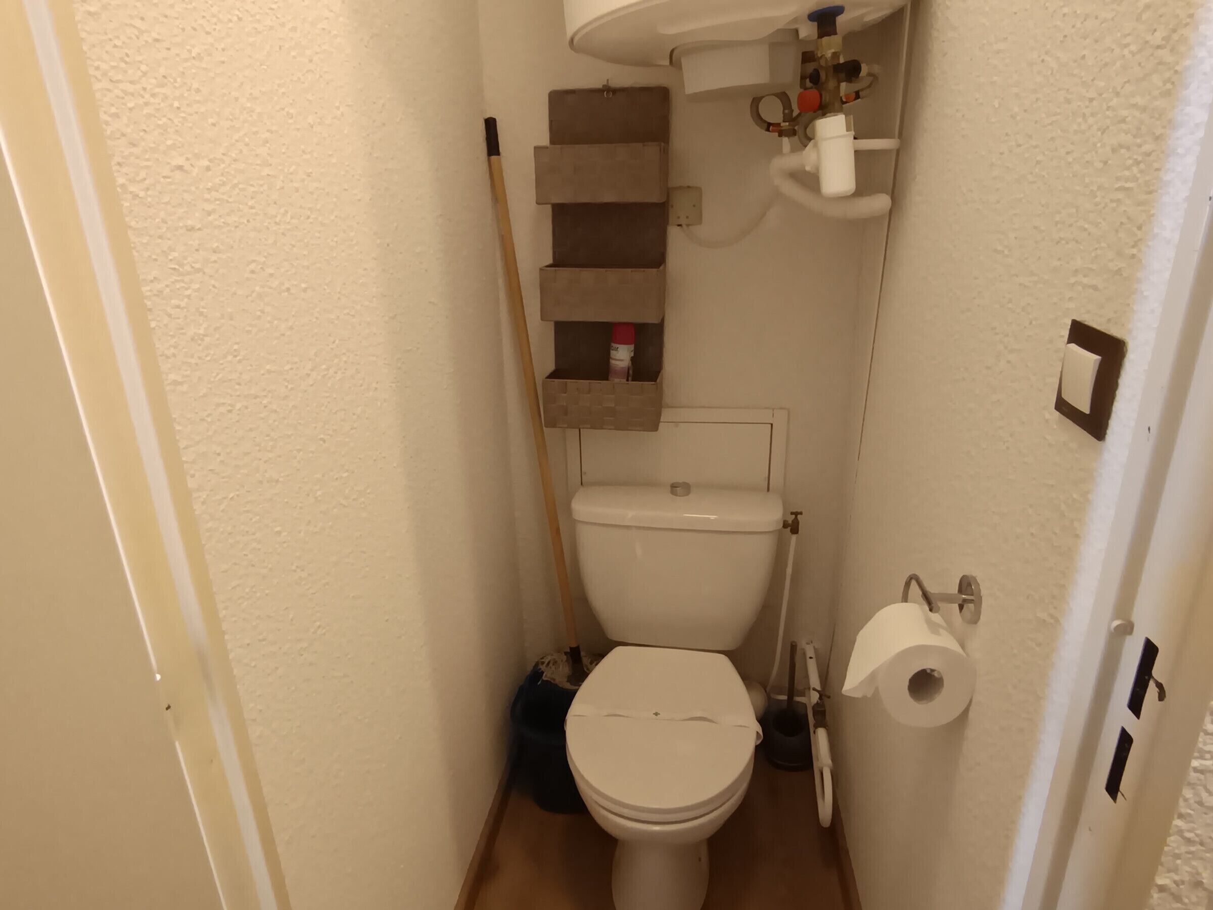 Pieni, käytännöllinen kylpyhuone, jossa on wc, hylly hygieniatuotteille ja seinää vasten nojaava luuta.