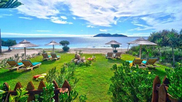 Un espace de détente aménagé au bord de la plage, avec des chaises longues et des parasols, une pelouse verdoyante et une vue sur la mer et une île lointaine.