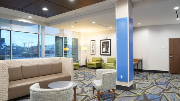 A hotel lobby with a sofa, chairs, a small table, a lamp, and framed artwork on the wall.