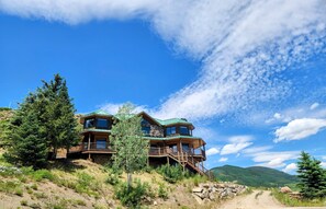 A two-story log cabin with a stone foundation, wooden deck, and multiple windows. - Luxurious Mt CB Home | 6 Bedroom | Best Views in CB | Hot Tub | Gas Grill (Mt. Crested Butte)