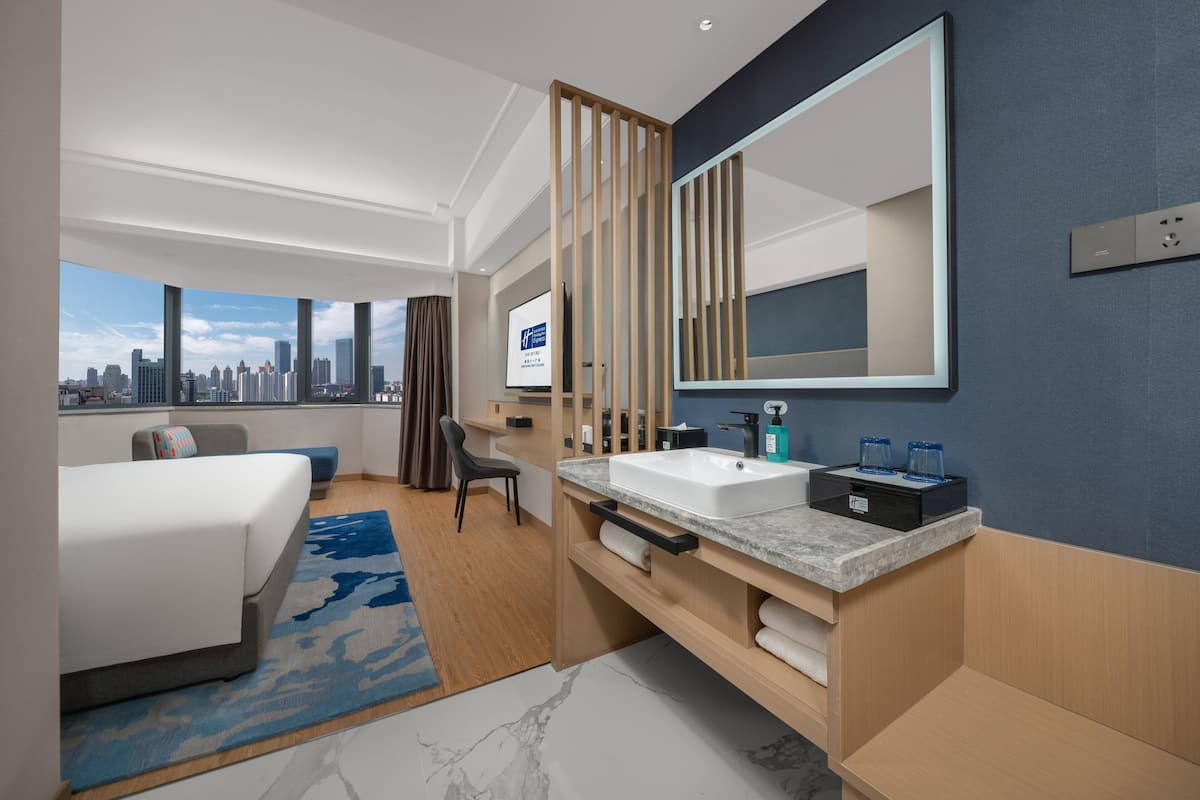 A modern hotel room with a large bed, a bathroom with a sink and mirror, and a city view.