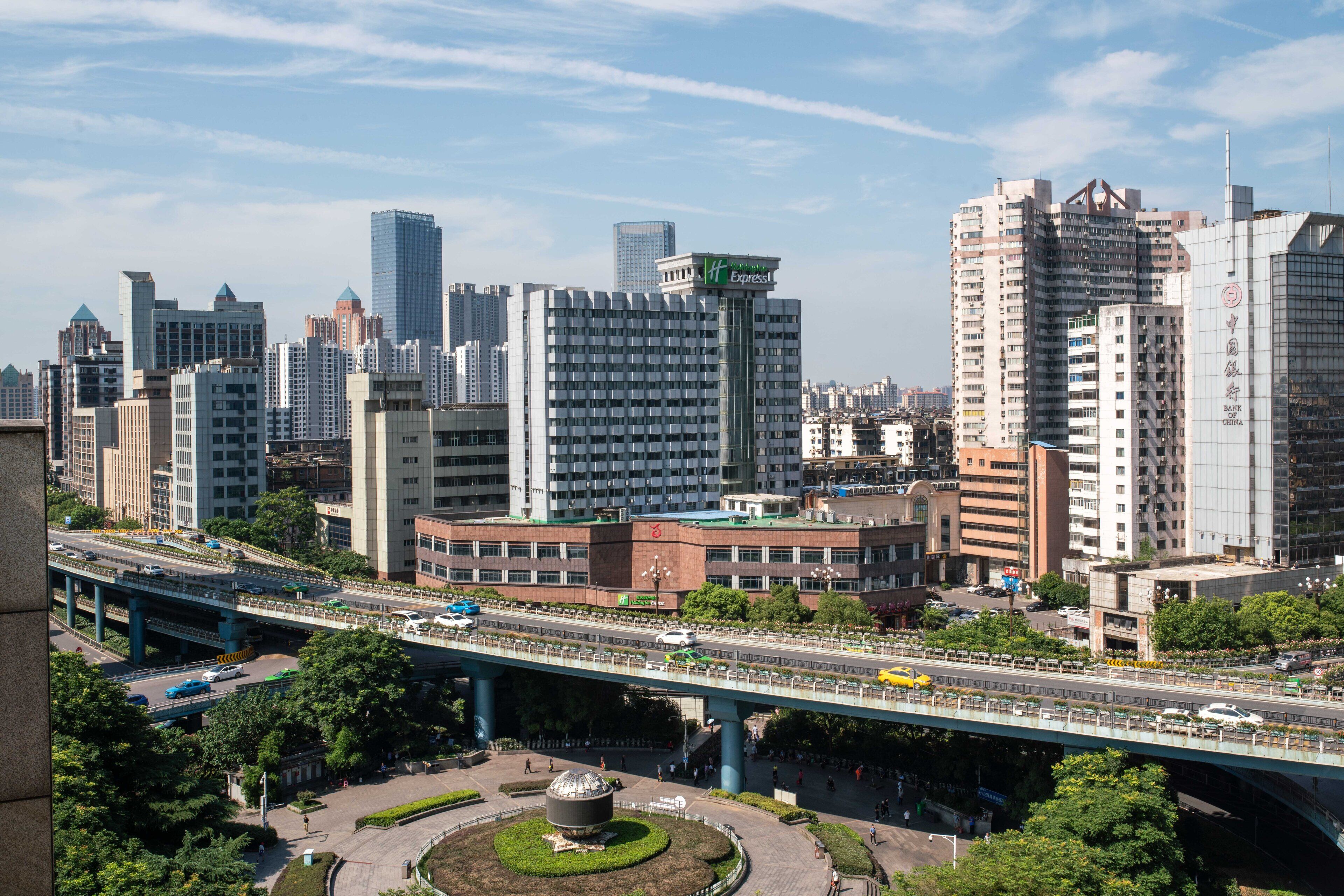 A cityscape with modern high-rise buildings, a prominent hotel with a green sign, and a busy overpass with vehicles.
