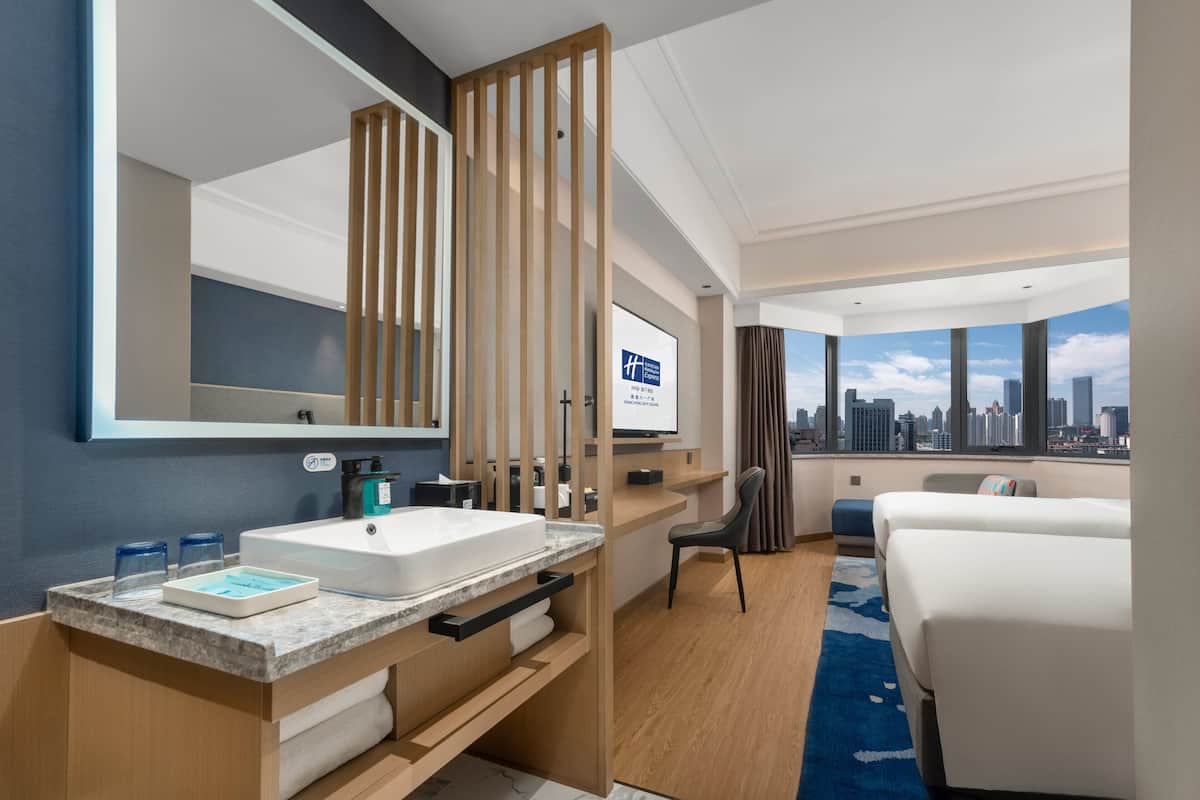 A modern hotel room with a large window offering a city view, a private balcony, a large mirror, a wooden partition, and a bed with a cityscape view.