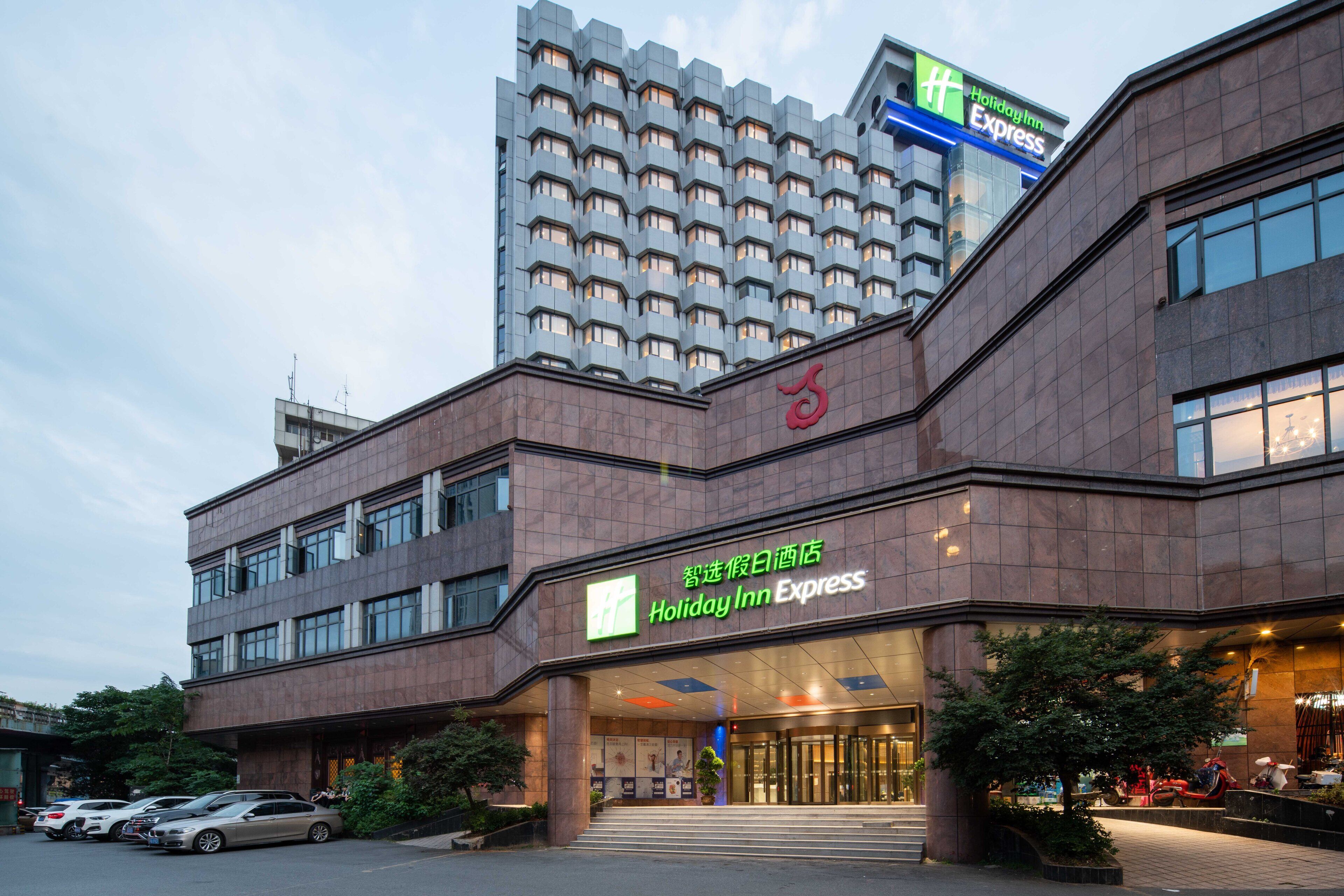Un bâtiment de la chaîne Holiday Inn Express, doté d’un design moderne, avec une entrée imposante et un parking où sont stationnées plusieurs voitures.